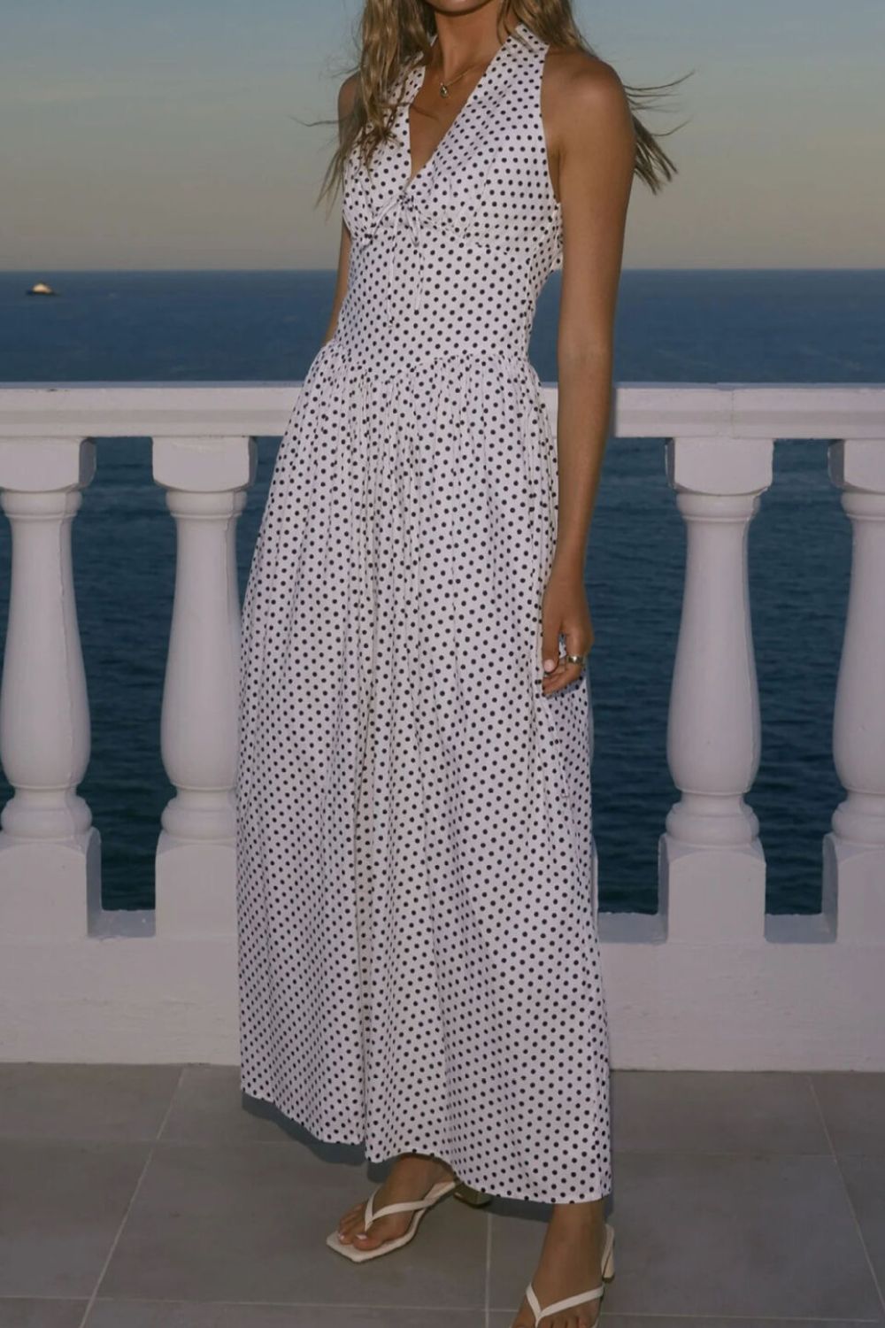 Woman in a white dress with black patterns standing by a railing overlooking the ocean.