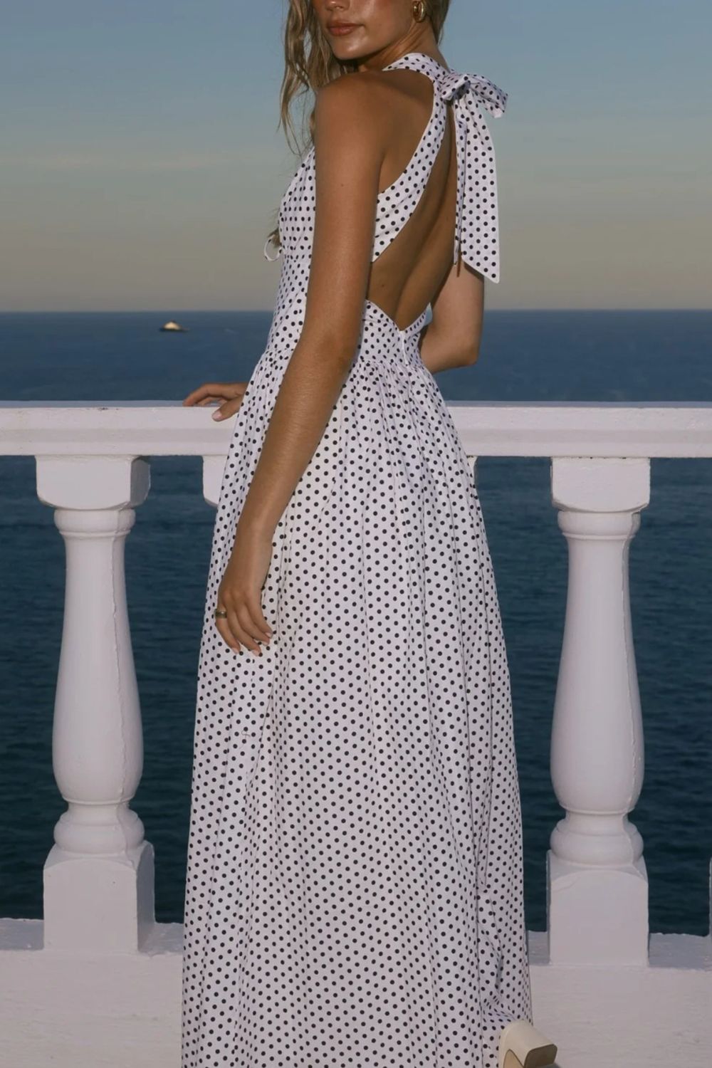 Woman in a polka dot dress with open back standing on a balcony overlooking the ocean.